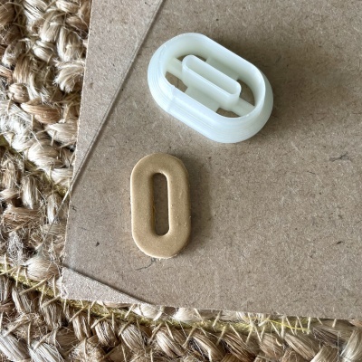 Dois acessórios ovais, branco de plástico e bege opaco, sobre cartão castanho e tapete de fibras naturais.