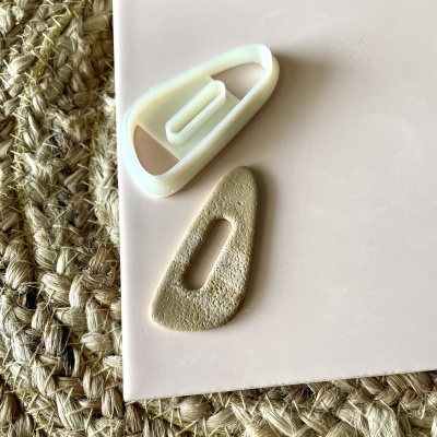 Dois grampos de cabelo, branco e dourado, sobre superfície rosa clara ao lado de tapete trançado