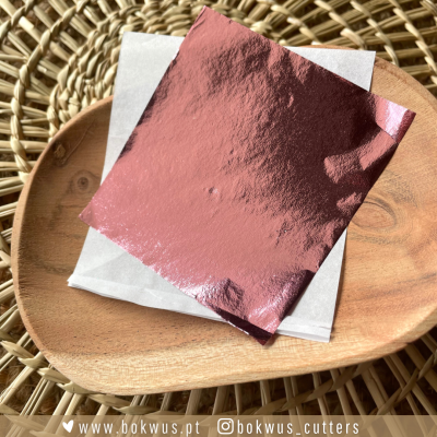 Pacote com papel metalizado rosa e papel branco sobre prato de madeira e superfície de palha com texto divulgativo.