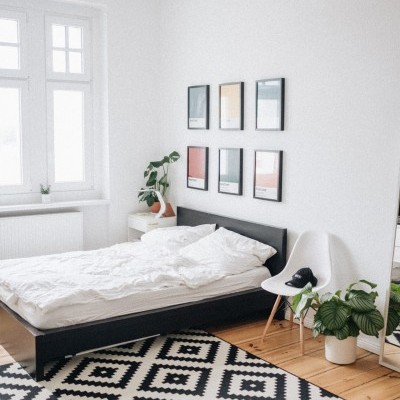 Quarto minimalista com cama preta, roupa de cama branca, tapete geométrico preto e branco e cadeira branca com planta