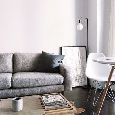 Sofá cinza com almofada escura numa sala com mesa de centro de madeira, cadeira branca, candeeiro de pé e quadro apoiado na parede.