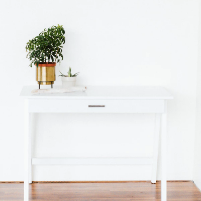 Mesa branca com duas plantas em vasos sobre ela, parede branca e chão de madeira