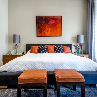 Quarto com cama de estrutura preta, almofadas laranja e preta, bancos vermelhos, mesas de cabeceira com lâmpadas, quadro abstrato e cortinas cinzentas