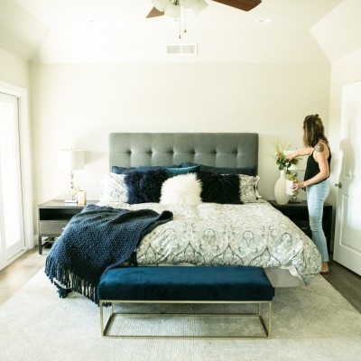 Quarto bem iluminado com cama decorada e mulher junto a mesa de cabeceira