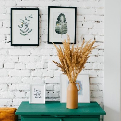 Cómoda verde com vaso de cerâmica castanha e plantas secas, molduras com folhas e placa com texto numa parede de tijolos branca