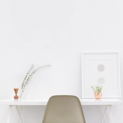 Mesa branca de trabalho com cadeira cinzenta, castiçal, vaso de cobre e quadro branco na parede