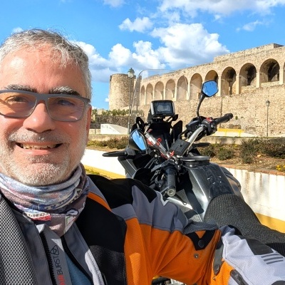 Homem com casaco de mota junto a mota preta em cenário histórico com torre de pedra