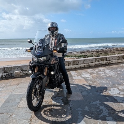 Motociclista numa moto preta junto à praia com céu e mar ao fundo