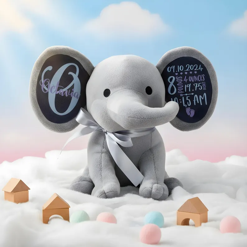 Pelúcia de elefante cinzento com texturas suaves e texto personalizado nas orelhas, rodeado de pequenas casas de madeira e bolas coloridas sobre tecido branco