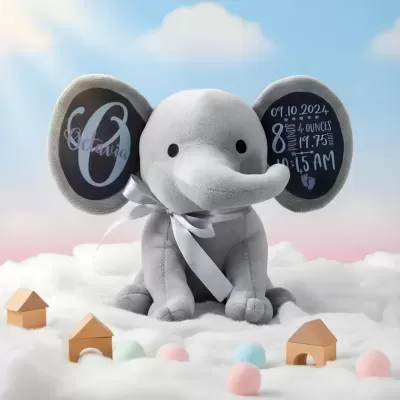 Pelúcia de elefante cinzento com texturas suaves e texto personalizado nas orelhas, rodeado de pequenas casas de madeira e bolas coloridas sobre tecido branco