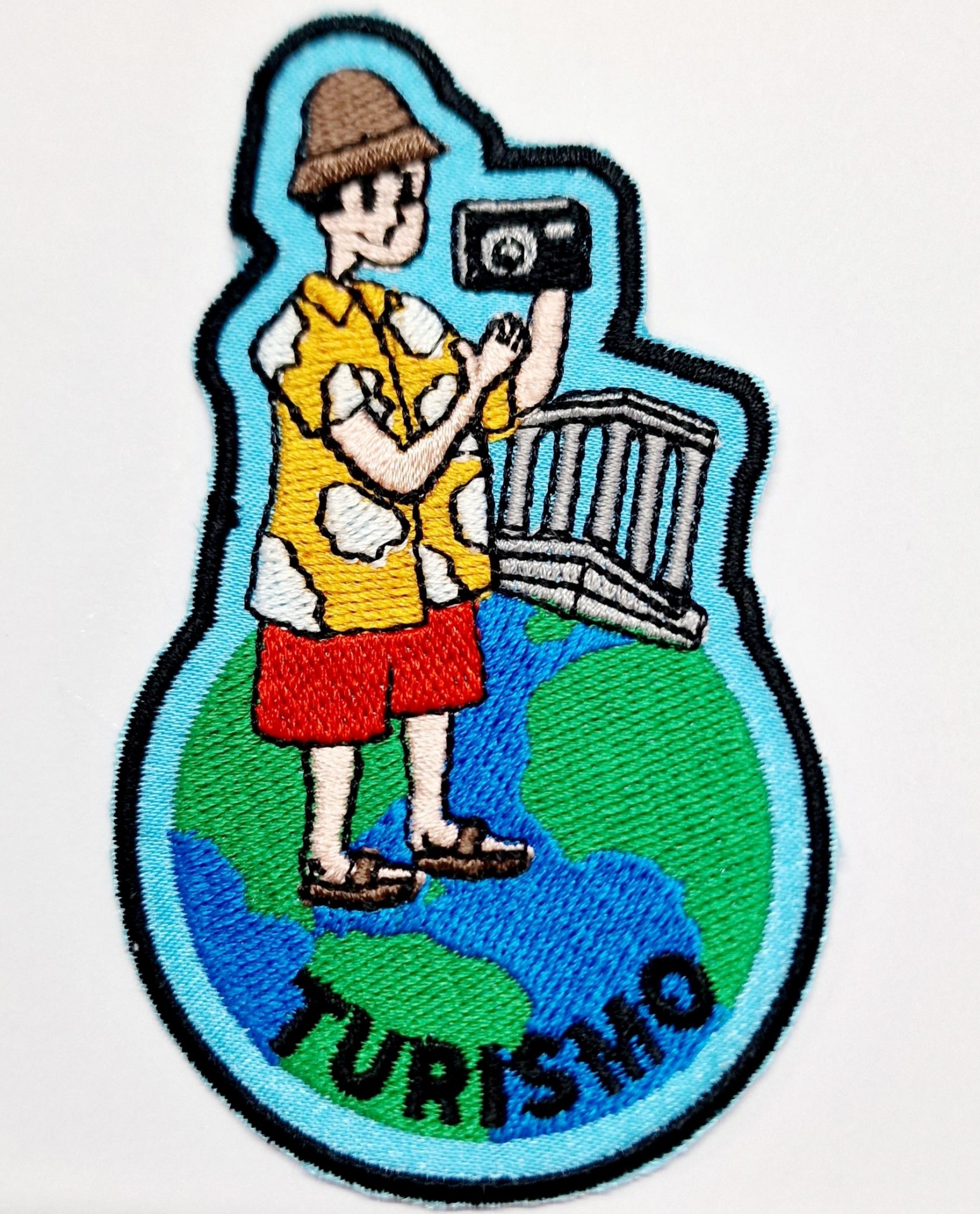 Emblema bordado representando um turista vestido com roupa colorida, sobre um globo terrestre com inscrição TURISMO.