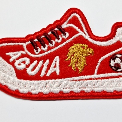 Emblema bordado em forma de sapatilha vermelha com detalhes em branco, cordões pretos e decoração de águia e bola de futebol