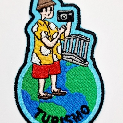 Emblema bordado representando um turista vestido com roupa colorida, sobre um globo terrestre com inscrição TURISMO.