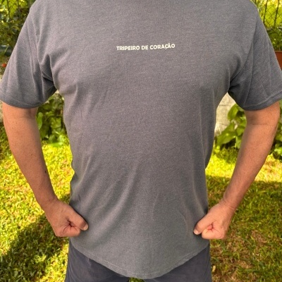 T-shirt cinza com texto branco TRIPEIRO DE CORAÇÃO ao centro, calções escuros, numa área exterior com relva e cerca com folhas verdes.