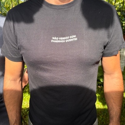 T-shirt preta com texto branco NÃO VENHAS COM PANINHOS QUENTES vestida por homem ao ar livre