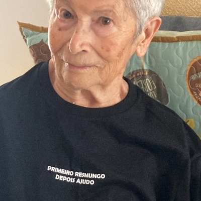 Mulher idosa com t-shirt preta com texto branco sentada em cadeira com almofadas coloridas
