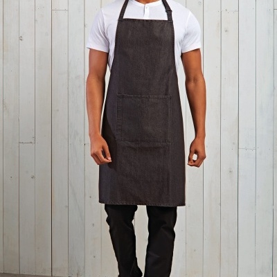 Homem a usar avental preto de tecido denim com bolso frontal, t-shirt branca, calças pretas e ténis brancos em fundo de madeira branca.