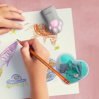 Mãos a desenhar com lápis colorido, apontador cinzento em forma de pata, caixa de clips verde e desenhos coloridos sobre papel branco.