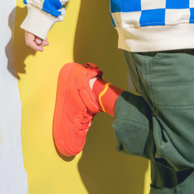 Pessoa com casaco quadriculado azul e branco, calças verdes e sapatilhas laranja contra parede amarela