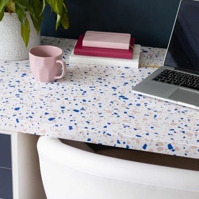 Mesa de trabalho com tampo branco com padrão terrazzo, caneca rosa, caixas, laptop e planta