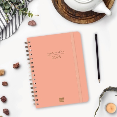 Agenda 2026 cor de rosa com elástico e espiral ao lado de caneta preta e chávena de chá