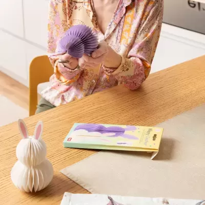 Decoração de Páscoa de papel em forma de coelho lilás e branco sobre mesa de madeira.