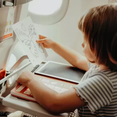 Criança a desenhar num avião com livro de atividades e folha com desenho de pássaro