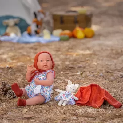 Boneca de bebé com roupa floral e acessórios vermelhos com boneco catinho e cardigan vermelho ao lado, em chão de terra e pinhas.