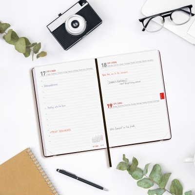 Mesa branca com agenda aberta, câmara fotográfica, caderno, óculos, caneta e folhas verdes