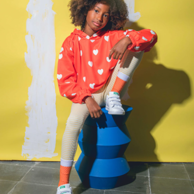 Criança com camisola vermelha de corações, leggings claras, meias laranjas e ténis brancos sentada em objeto azul