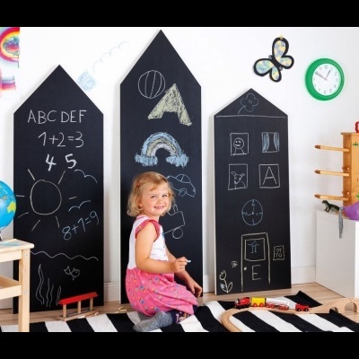 Painéis de ardósia em forma de casa com desenhos, menina a desenhar no chão, globo terrestre, relógio e prateleira com livros.