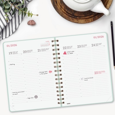 Agenda aberta com capa verde claro, anotações, caneta preta, pano listado, pedra rosa, chaleira branca com suporte de madeira sobre mesa branca