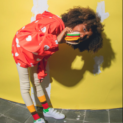 Criança com casaco vermelho de corações, calças riscas e meias coloridas, segurando hambúrguer de feltro.