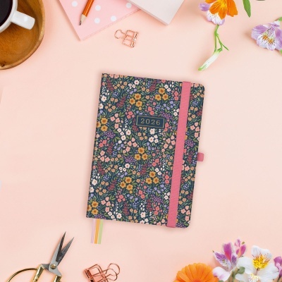 Agenda 2026 floral com elástico rosa entre objetos de escritório e flores num fundo rosa claro