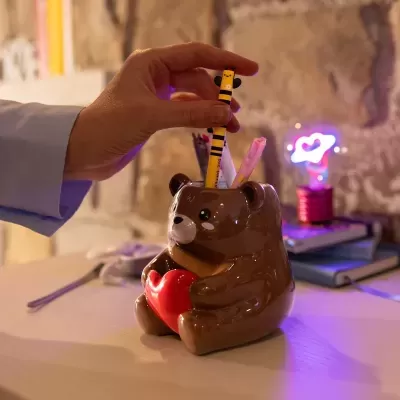 Porta-canetas de cerâmica em forma de urso castanho segurando coração vermelho com canetas coloridas