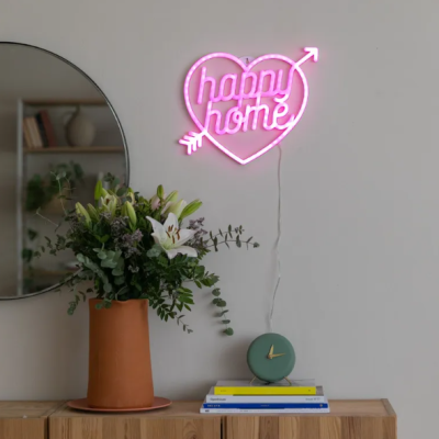 Neon decorativo rosa com texto happy home em coração, vaso com flores, relógio e livros sobre cómoda junto a espelho redondo