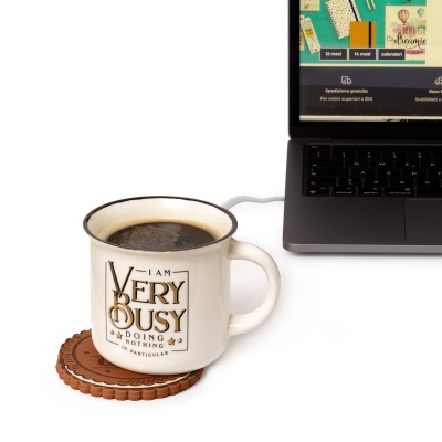 Caneca branca de cerâmica com texto e café, sobre suporte castanho, próximo de portátil cinzento.