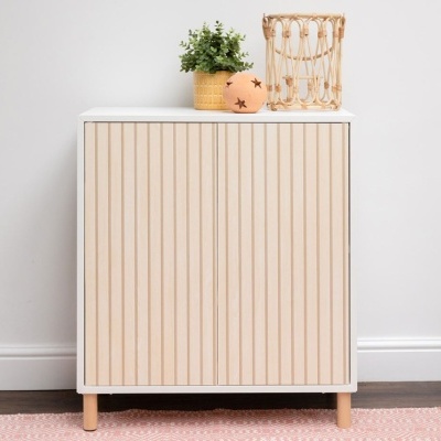 Móvel de madeira clara com ripas verticais sobre tapete rosa e decoração minimalista.