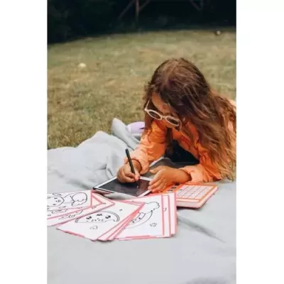 Criança desenha numa tablet ao ar livre rodeada de desenhos em folhas e um tablet com teclado