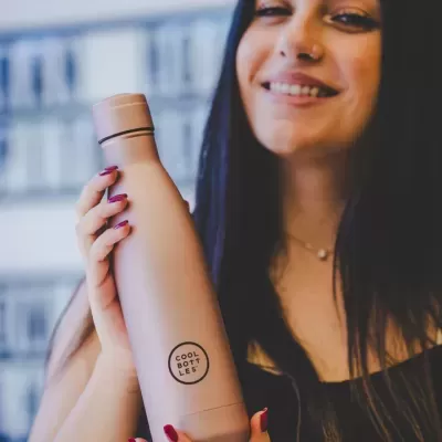 Garrafa térmica bege com logo COOL BOTTLES sendo segurada por uma mulher.