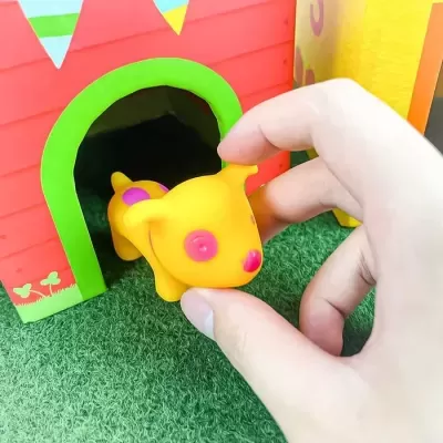 Boneco amarelo e rosa em forma de cão a ser segurado por uma mão junto a uma casinha de brinquedo vermelha e verde sobre relva artificial
