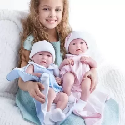 Menina com duas bonecas realistas vestidas de azul e rosa sentada num sofá branco