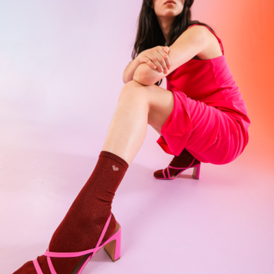 Mulher sentada com vestido rosa e sandálias rosa de salto, usando meias castanho-avermelhadas com padrão de coração