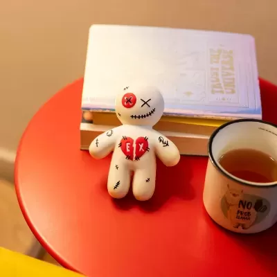 Boneco de vinil com botão vermelho no olho e coração partido colocado numa mesa vermelha junto a livros e uma caneca