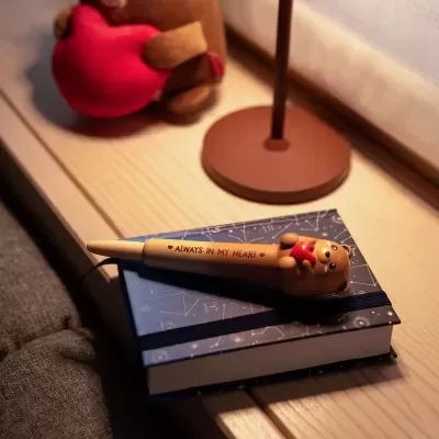 Caneta com tampa de ursinho sostendo coração vermelho sobre caderno azul com tema de constelações