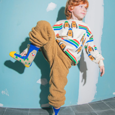 Criança com camisola branca com padrão de cães e riscas coloridas, calças castanhas peluche e meias azuis coloridas em parede azul