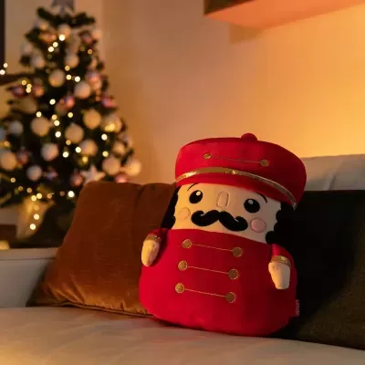 Almofada de peluche vermelha em forma de soldado com botões dourados no sofá, com árvore de Natal desfocada ao fundo