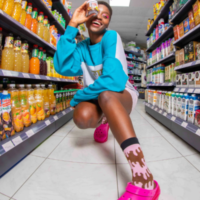 Mulher agachada em corredor de supermercado com frasco pequeno, vestindo camisola azul, meias e chinelos rosa