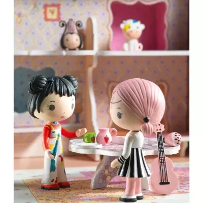 Duas bonecas miniatura numa mesa com chávenas, jarra e guitarra num cenário de casa de brinquedo.