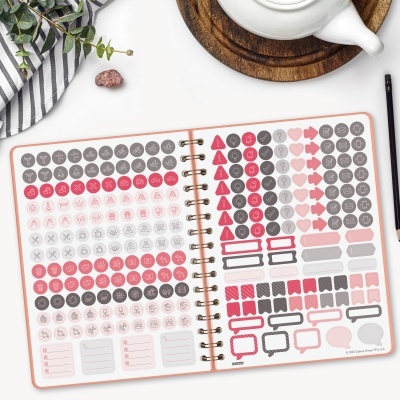 Caderno com autocolantes organizadores coloridos aberto numa mesa com caneta, planta e bule.